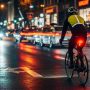 A cyclist in a bustling city street at night, illuminated by bright front and rear bike lights, highlighting urban visibility gear.