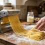 Electric pasta machine extruding fresh spaghetti strands onto a floured wooden kitchen surface