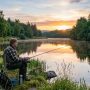 A person sitting peacefully on a riverbank fishing at sunrise surrounded by green trees and calm water